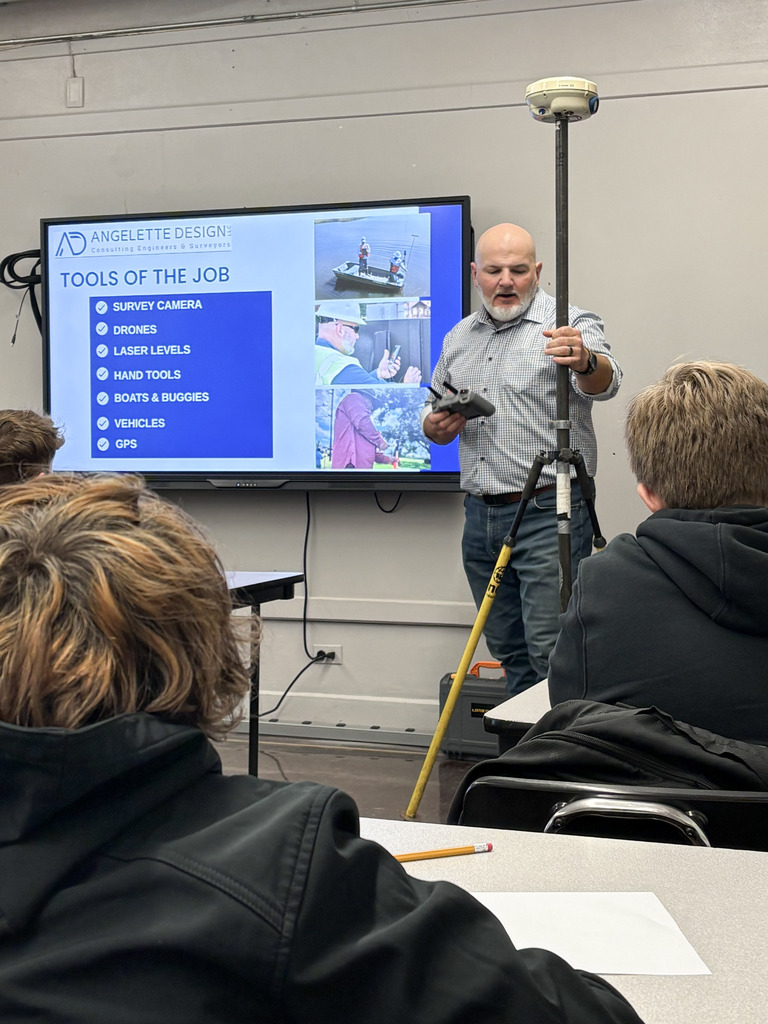 A guest speaker from Angelette Design stands beside a tall survey GPS trip pole and holds a remote device while presenting "Tools of the Job" on a large digital screen to a room of middle school students.