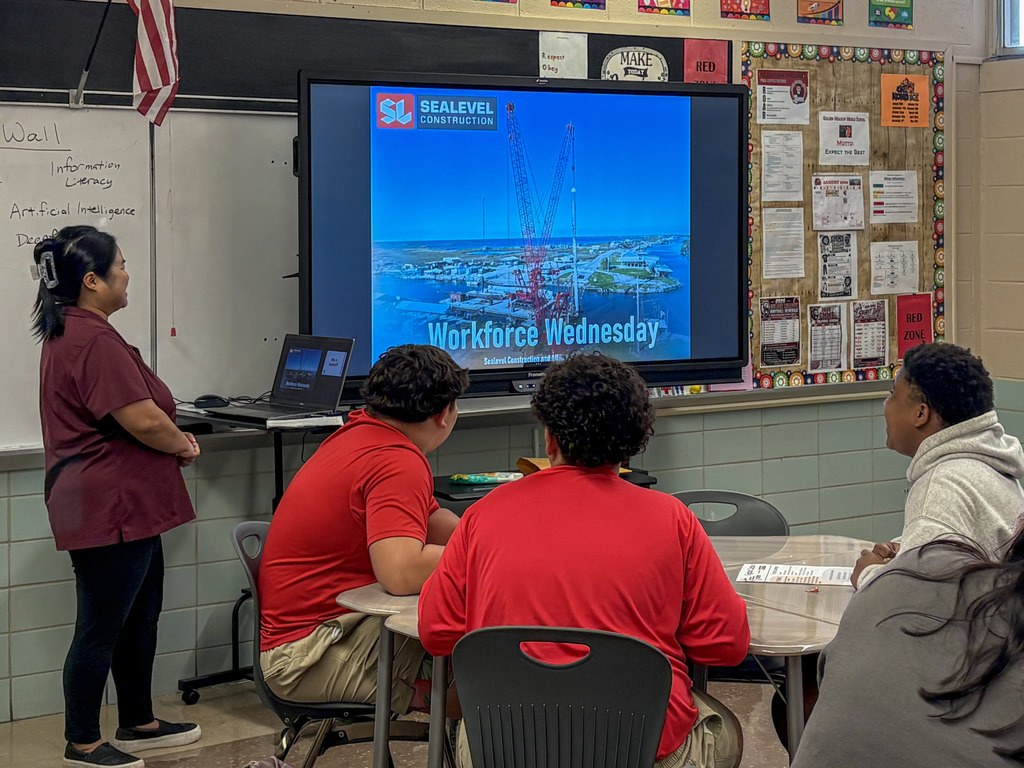 A guest speaker stands in front of a digital screen presenting a photo of construction cranes and the text "Workforce Wednesday," while a group of students sits around a table in the foreground, watching the presentation.