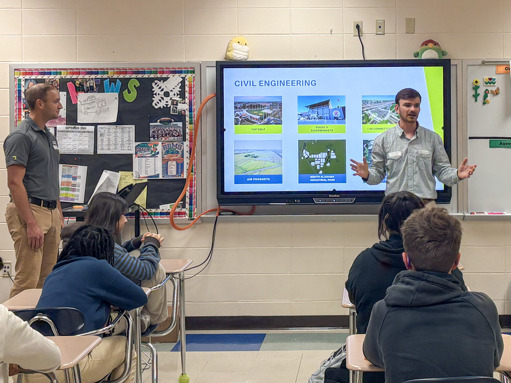 wo guest speakers stand next to a large digital monitor displaying a slide titled "Civil Engineering" with photo examples of local infrastructure projects, speaking to a class of attentive middle school students.