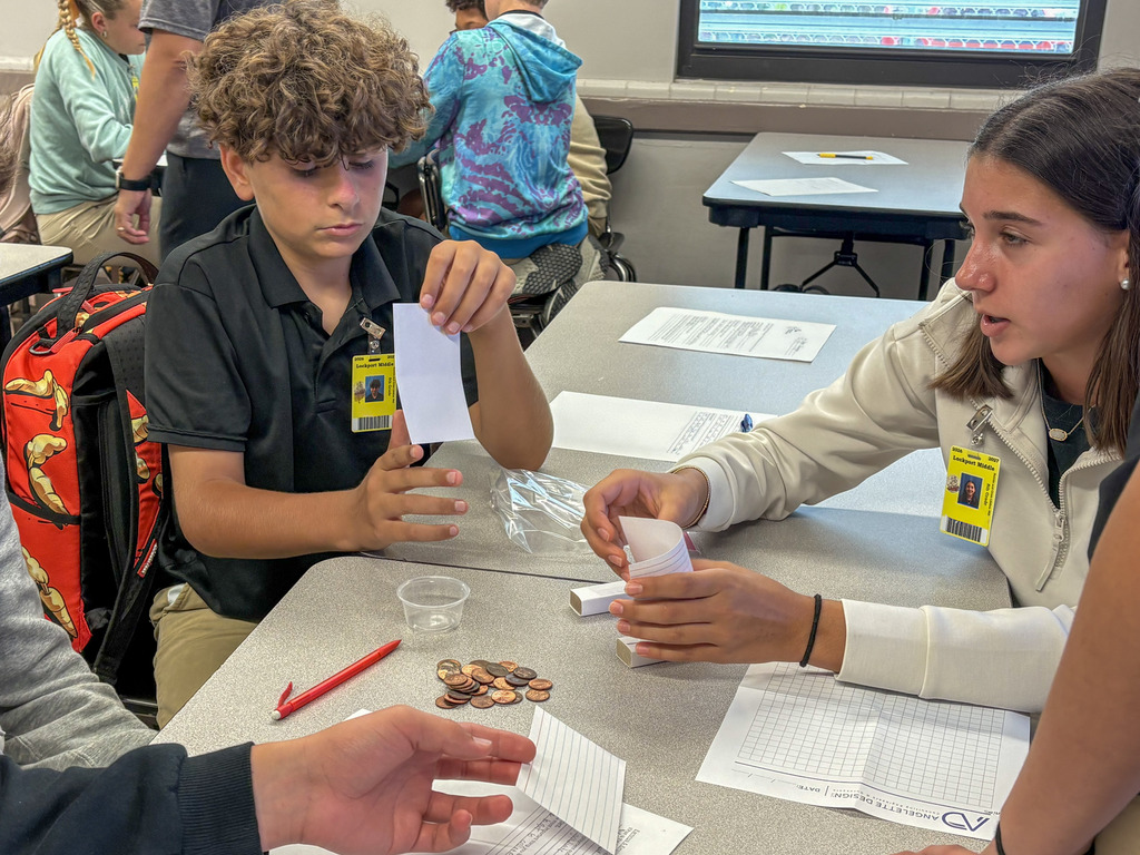 Close-up of middle school students sitting at classroom tables participating in a hands-on financial or math activity with paper slips, pencils, and small piles of pennies.