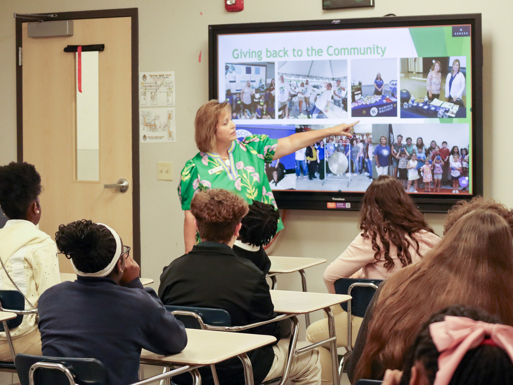 A guest presenter in a green floral blouse points to a slide titled "Giving back to the Community" on a digital display showing photos of Hancock Whitney bank community events to a room of middle school students.
