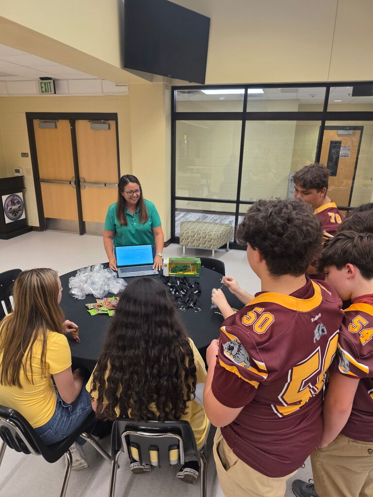 People gather around a table with a laptop. Two are wearing maroon jerseys.
