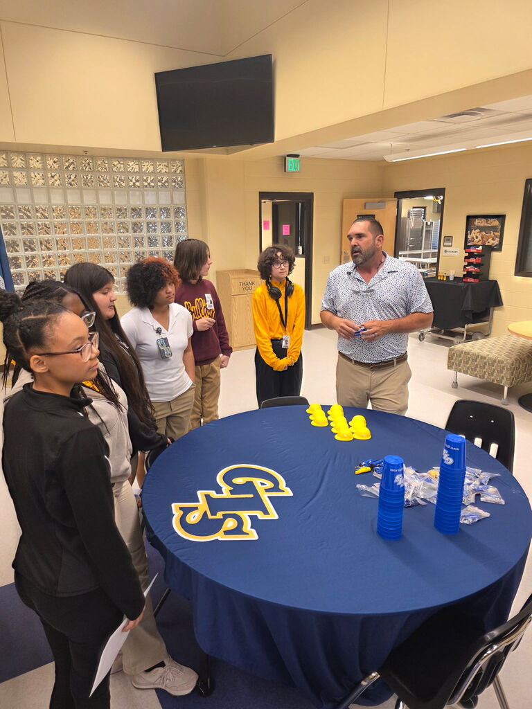 A group of students and an adult male in a classroom, standing around a table covered with blue cloth.