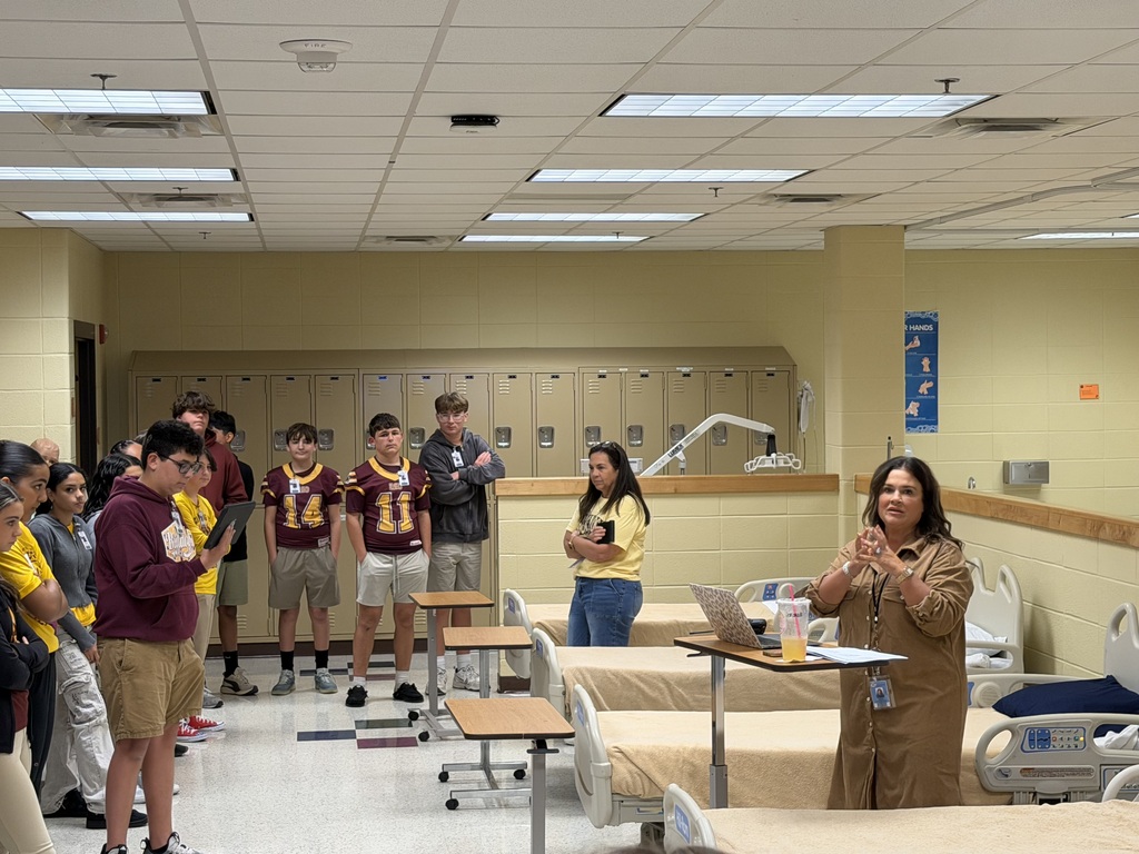 Group of students in a classroom setting with lockers, tables, and a person with a laptop.