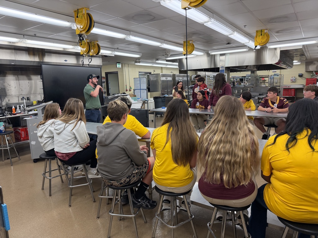 A group of students in yellow shirts seated around tables in a classroom, listening to a person standing at the front.