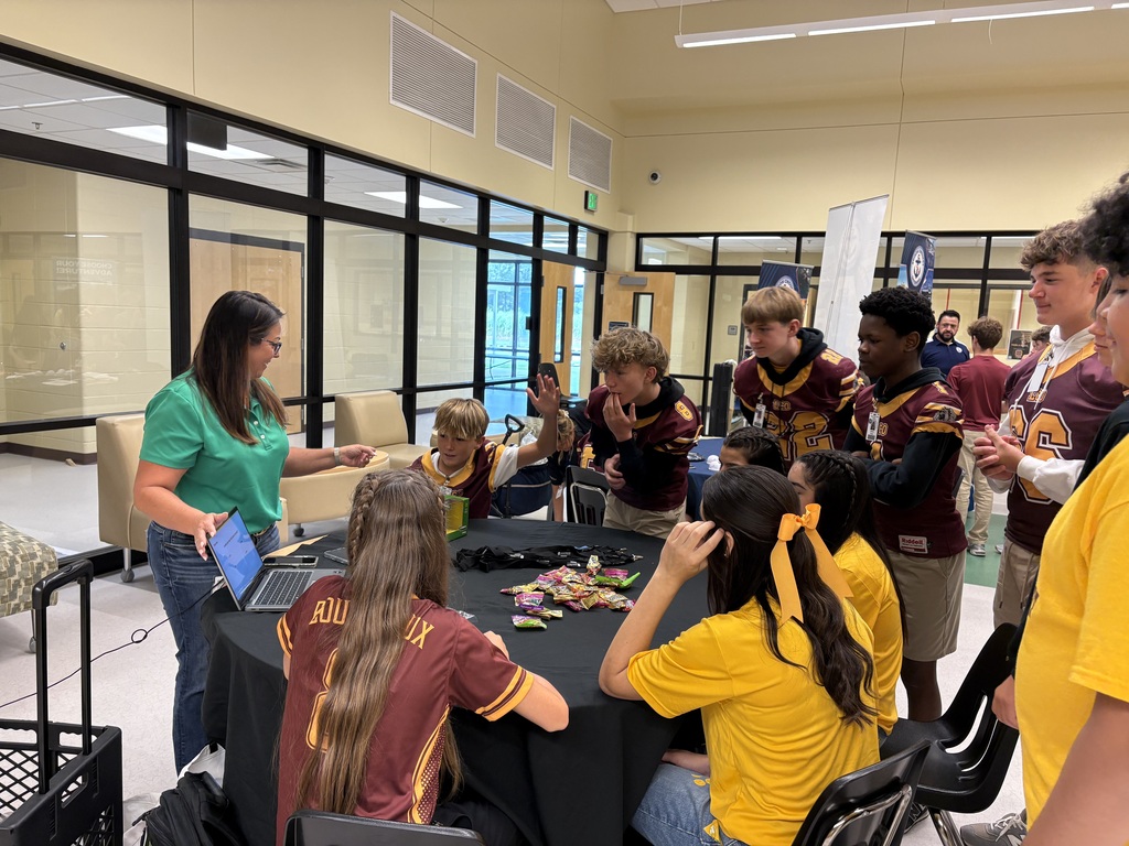 A group of students in yellow and maroon shirts gather around a table with a laptop.