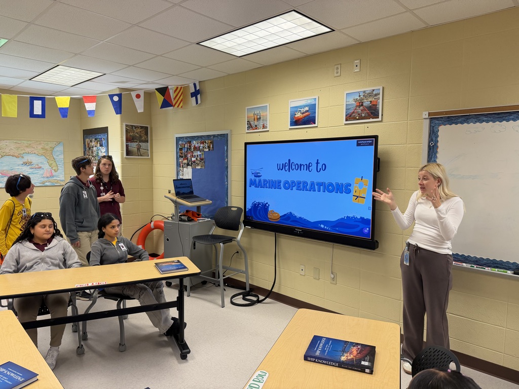A woman stands before a large monitor in a classroom. Students sit at tables. Flags and pictures adorn the yellow walls.