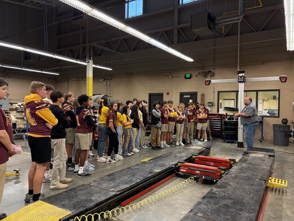 A group of students wearing yellow and maroon uniforms stands in a garage with a vehicle lift.