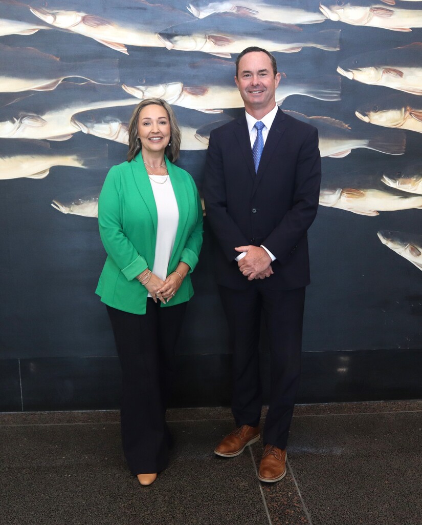 A man and a woman stand close together in front of a large fish mural on a wall.