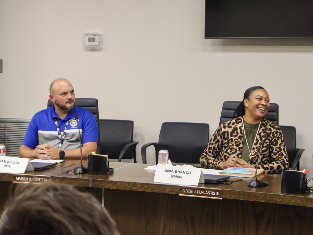Two individuals seated at a table, smiling, with name tags and microphones, and a television on the wall.