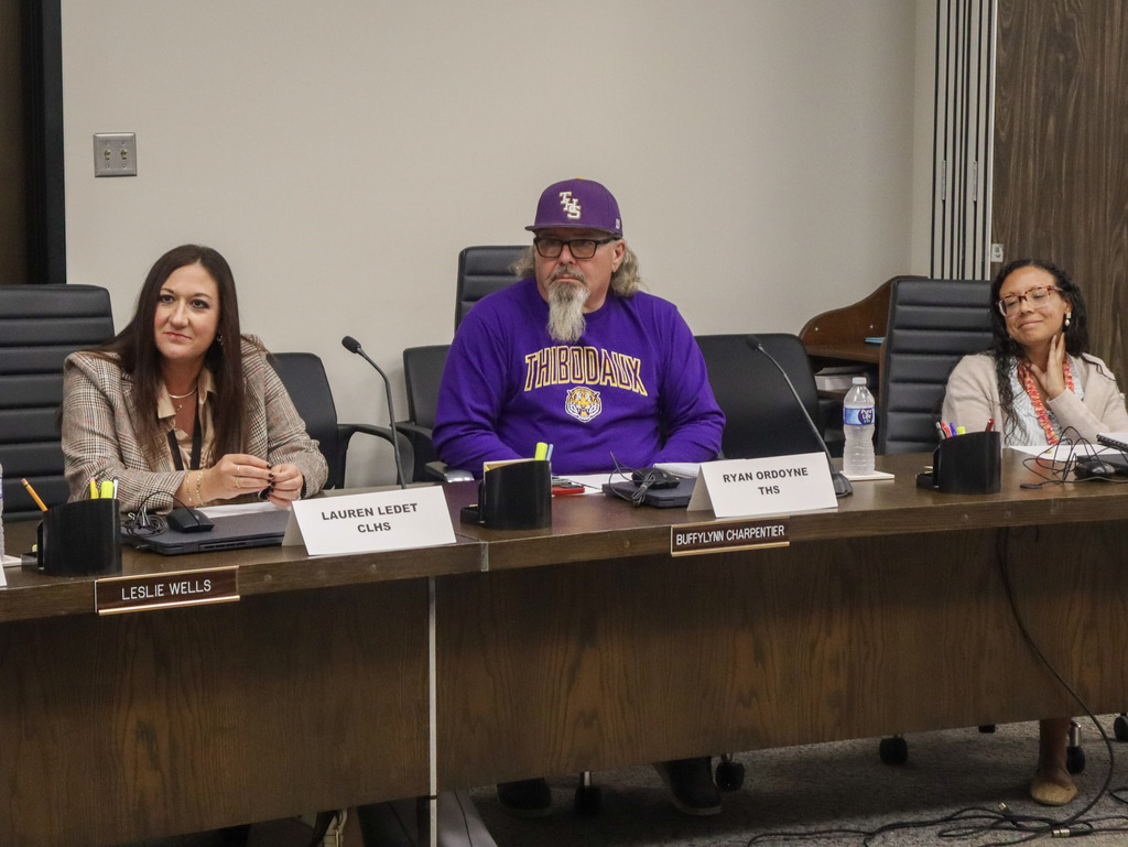 Three people seated at a long table, with nameplates, microphones, pens, and a water bottle in front of them.