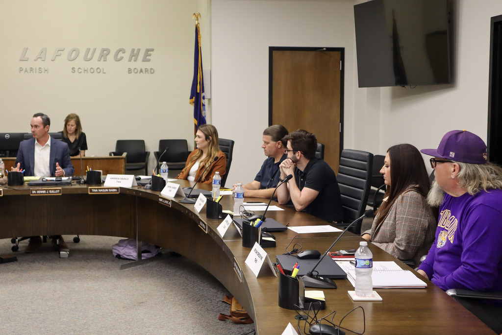 School board meeting with six adults seated around a table, three in suits, one with a purple hat.