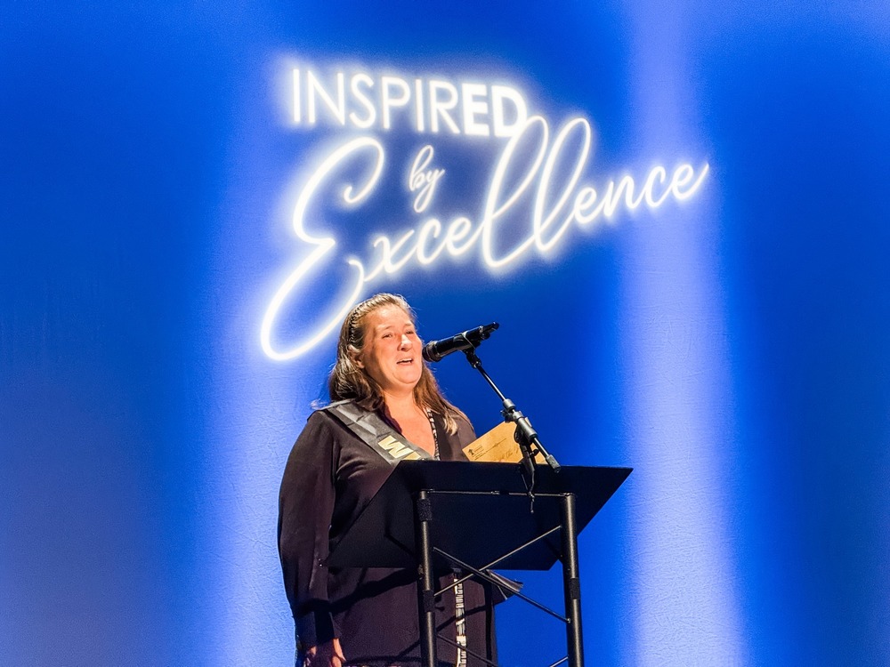 Michelle Deesi stands at podium with illuminated signage that reads, "Inspired by Excellence" overhead