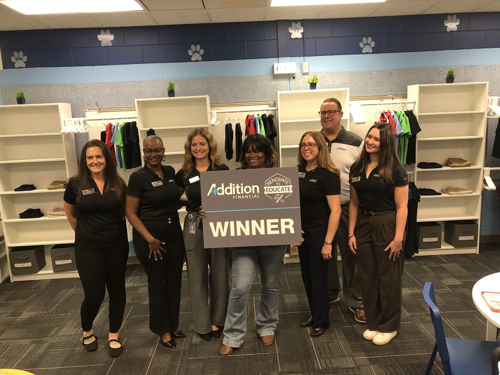 Eustis Heights Principal holding sign that says Addition Financial Renovate To Educate Winner. Other staff members are standing with her, all wearing black polo shirts