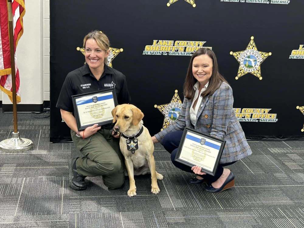 Canine handler Erica Stamborski and Florida Attorney General Ashley Moody kneeling beside vape dog Samba and holding certificates.