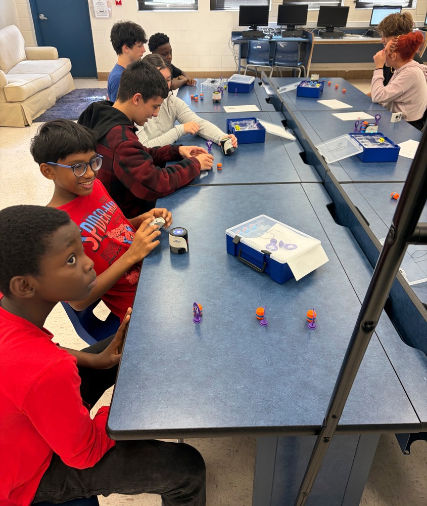 Students gather around tables in a classroom for a hands-on coding activity using small VEX robots and colorful coding pieces. Several students interact with the robots and materials while working together, with computer workstations visible along the back wall.