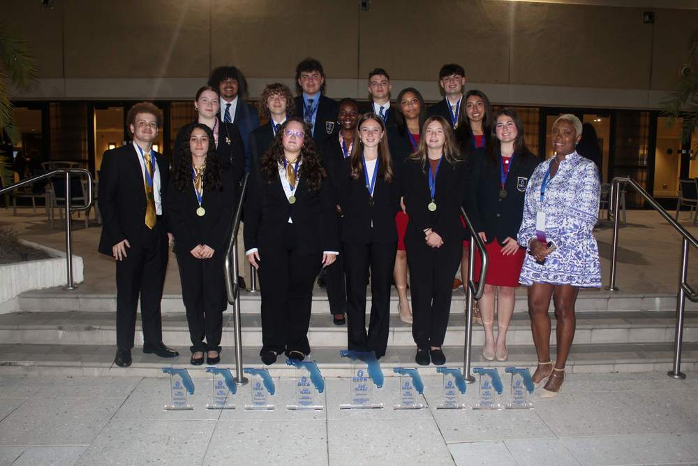 High School students in dark pants and blazers wearing medallions. Plaques at their feet. Teacher standing with them.