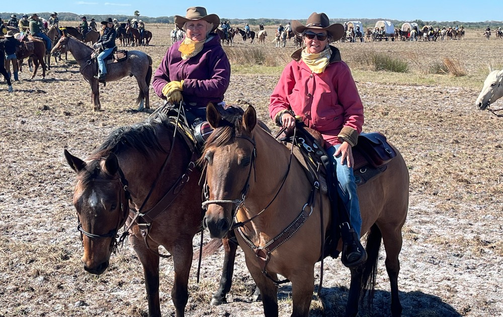 Two women on horses in open field wearing cowboy hats with lots of horses and riders in the background