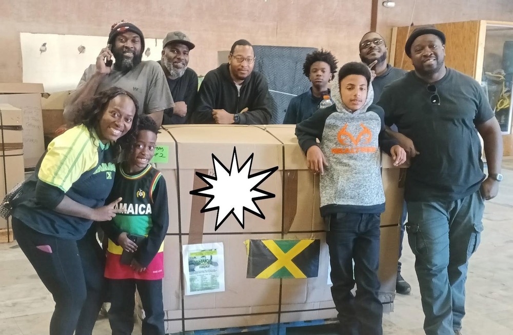 6 adults and 3 children standing around boxed items with a flag of Jamaica taped to the boxes.
