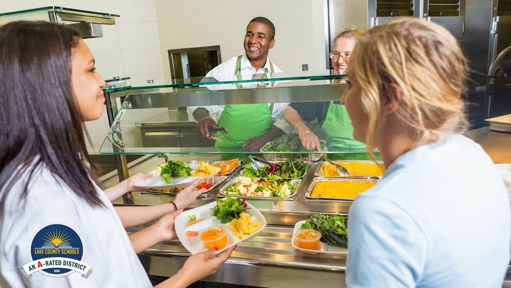 Teen girls get food in cafeteria line, served by a man and a woman, both smiling and wearing green aprons.