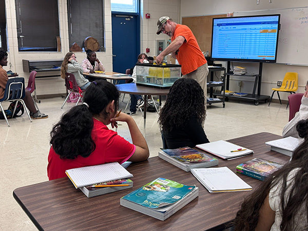 Mr. Bryan Russ demonstrates sound waves to students in his Oak Park Middle School science classroom. Students watch as he uses a large yellow bell submerged in a clear tank of water to make the effects of sound-producing vibrations visible. Students are seated around classroom tables with science textbooks and notebooks as they observe the hands-on demonstration.