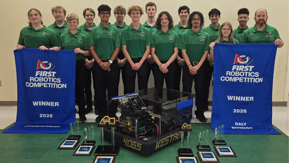 Team of students in green polo shirts posed with robot, six trophies, and two banners that say FIRST Robotics Competition Winne r 2026