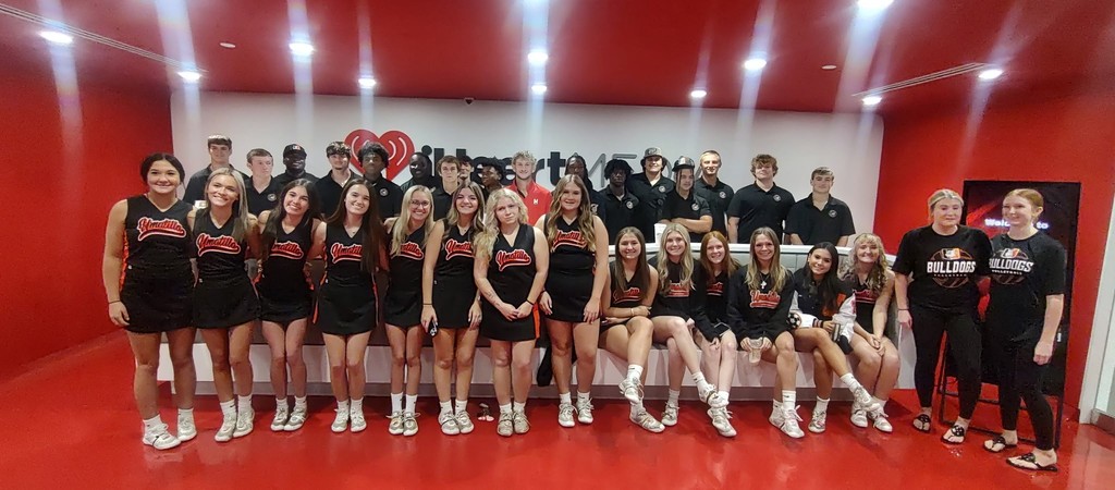 Umatilla High cheerleaders and athletes pose at iHeartRadio in front of the Johnny’s House logo, proudly representing the Bulldogs in orange and black.
