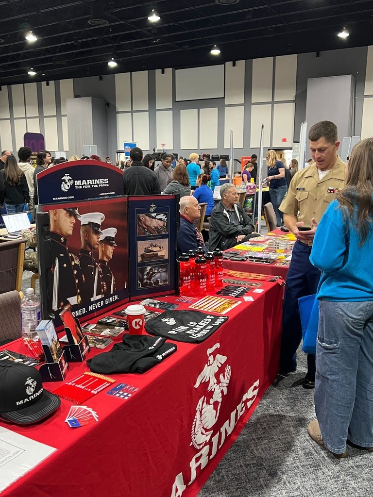 Female student speaking with a man in a Marine uniform in front of a table with a red Marines tablecloth topped with Marines posters, hats, T-shirts and other paraphernalia