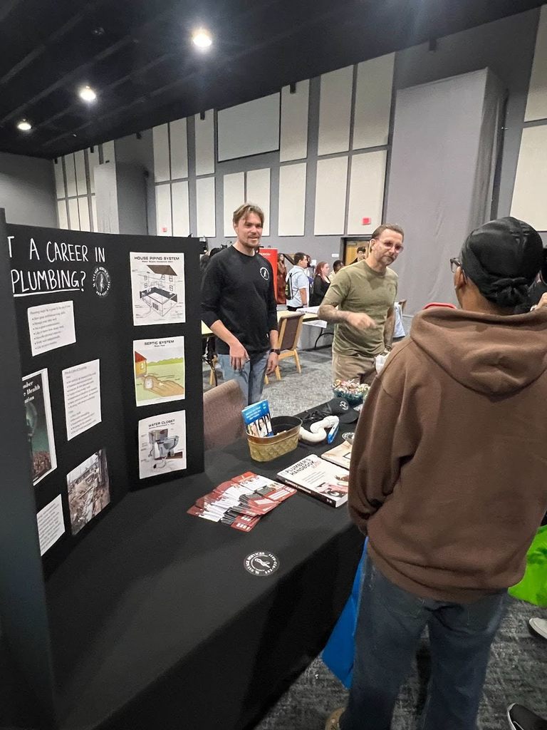 Male student talking to two male vendors standing behind a table with a display board that says "Want a Career in Plumbing?"