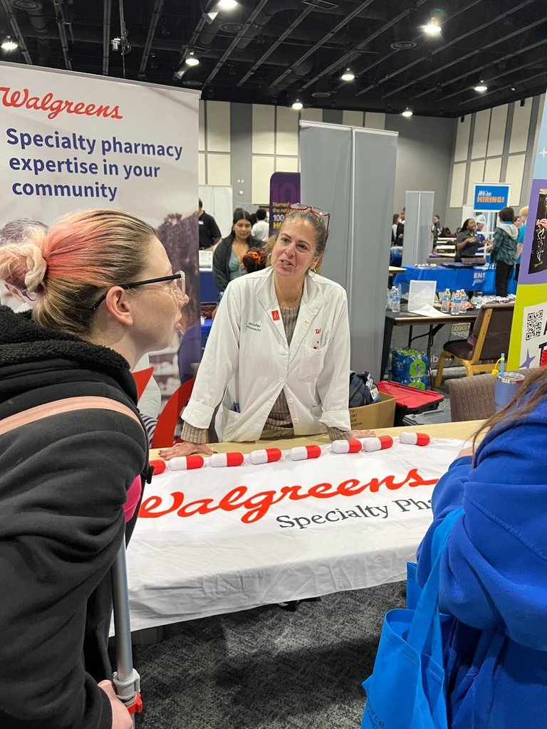 Female student talking with vendor in a lab coat standing behind a table with a Walgreen's Specialty Pharmacy tablecloth and a Walgreen's banner behind her. 