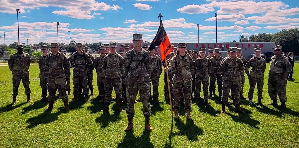 A group of Umatilla High School JROTC cadets in camouflage uniforms stand in formation on a grassy field under a bright blue sky with scattered clouds. Two cadets in the front hold a black and orange unit flag, and a building labeled “Falcon Field” is visible in the background.