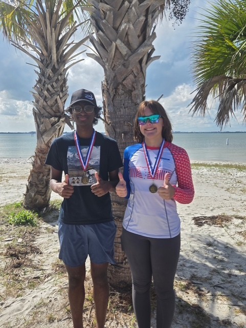 Two people stand on a sandy beach in front of palm trees, both smiling and giving thumbs-up gestures. They wear medals around their necks, suggesting a competition or award event. The ocean and a bright, partly cloudy sky are visible in the background.