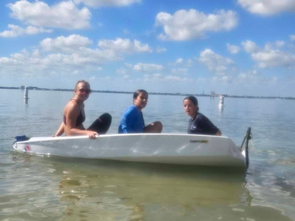 Three people sit in a small white sailboat floating on calm, shallow water. The sky is blue with scattered clouds, and distant shoreline structures are visible on the horizon. The group appears relaxed and enjoying a sunny day on the water.