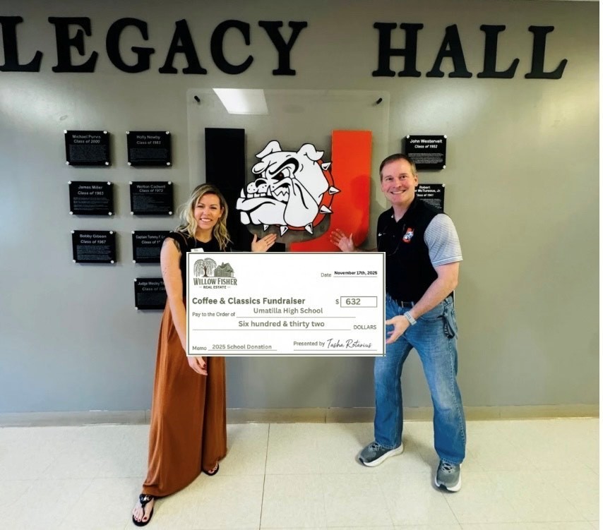 Two adults stand in the Umatilla High School Legacy Hall, smiling and holding an oversized check from Willow Fisher Real Estate. The check, made out to Umatilla High School, is for $632 from the “Coffee & Classics Fundraiser.” The school’s large Bulldog logo is displayed on the wall behind them along with plaques honoring past graduates.