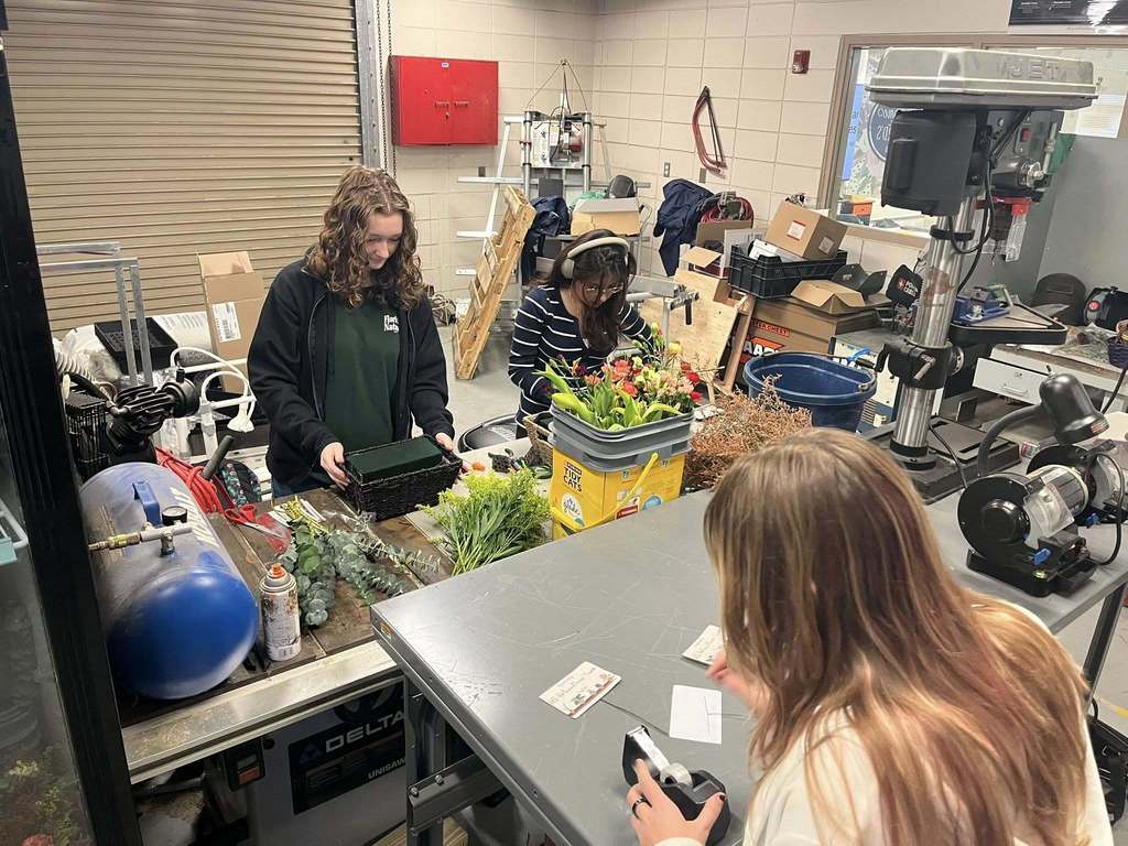 Three women work in an industrial workshop space on a project involving flowers and supplies. One woman with curly hair stands at a workbench holding a floral foam block. Another woman wearing headphones sits behind a table filled with colorful flowers and greenery. In the foreground, a third woman with light brown hair uses tape and labels at a gray table. The room is cluttered with tools, boxes, a drill press, and other equipment, creating a busy, hands-on workspace.