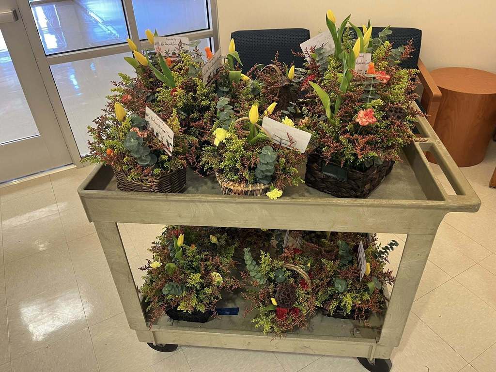 A rolling utility cart is filled with neatly arranged floral centerpieces in woven baskets. Each arrangement includes greenery, eucalyptus, reddish filler stems, and bright yellow tulips, along with small handwritten cards tucked among the flowers. The cart sits in a clean hallway near glass doors and a seating area, ready for delivery or display.