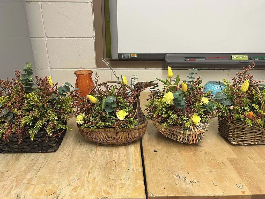Four woven baskets filled with colorful floral arrangements sit on a wooden table. Each basket contains greenery, yellow tulips, eucalyptus sprigs, and small yellow and orange flowers, with a mix of red-toned filler foliage. One basket in the center has a decorative handle shaped like a duck’s head. The background shows a classroom-style whiteboard and cinder block wall.