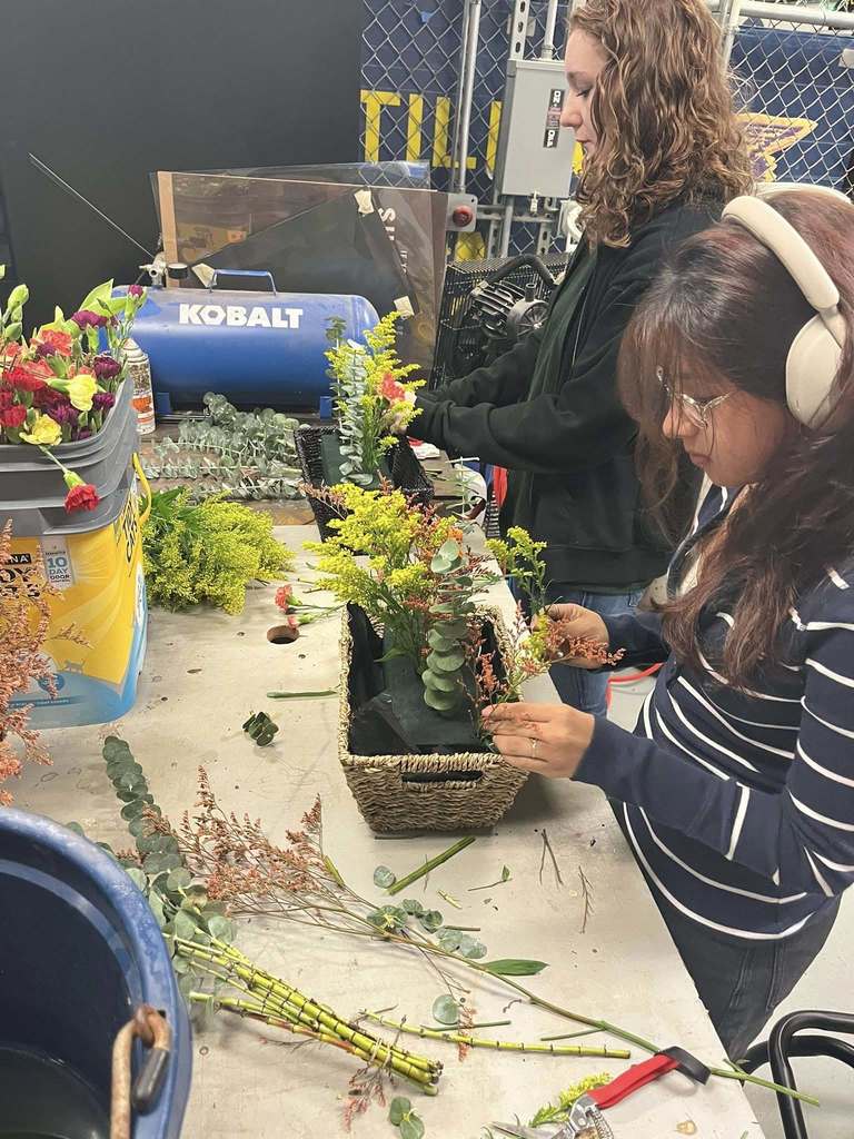 Two women work together at a large table arranging flowers. The woman in the foreground wears headphones and a striped sweater as she places stems into a rectangular woven basket filled with floral foam. The woman behind her, with curly hair and a dark jacket, arranges flowers in a separate container. The table is covered with assorted greenery, clippers, and loose stems, and the workspace has industrial shelving and equipment in the background.