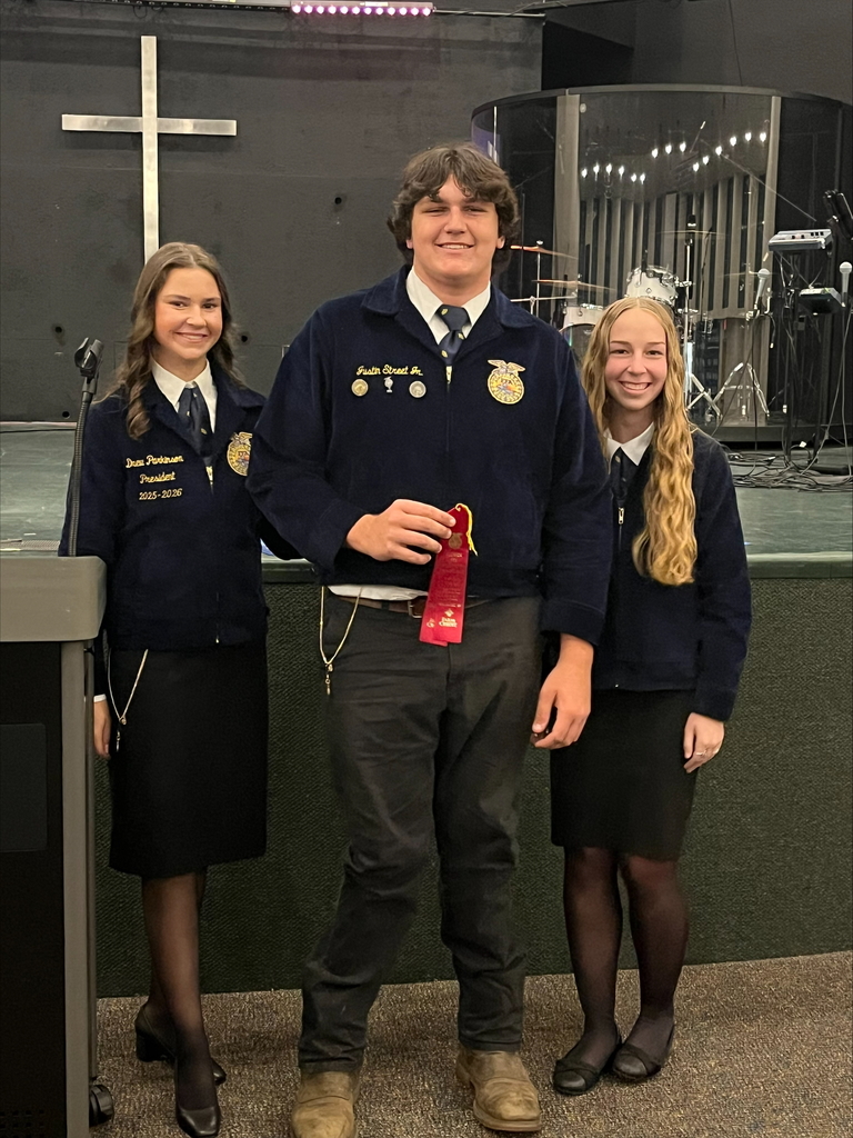 Three Umatilla FFA students stand together indoors, all wearing official blue FFA jackets, black skirts or pants, and black shoes. The student in the center, holding a red ribbon, stands slightly in front of the two students beside him. They are smiling, and behind them is a stage area with a large cross on the wall and musical instruments, including drums, visible in the background.