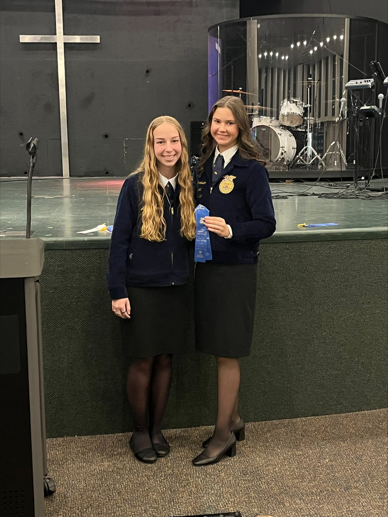 Two Umatilla FFA students stand together indoors, both wearing official blue FFA jackets and black skirts. One student holds a blue award ribbon as they smile for the photo. Behind them is a stage area with a large cross on the wall and a drum set enclosed in a transparent sound booth.
