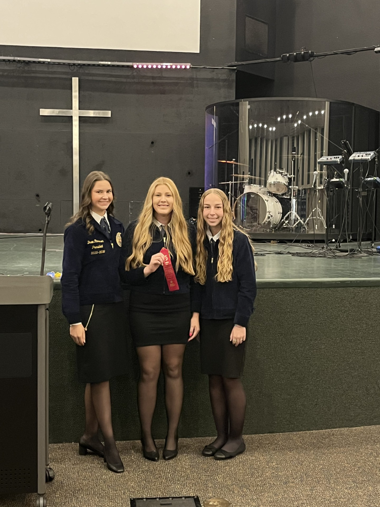 Three Umatilla FFA students stand together indoors wearing official blue FFA jackets and black skirts. The student in the center holds a red award ribbon while all three smile for the photo. Behind them is a stage area with a large cross on the wall and a drum set inside a transparent sound booth.
