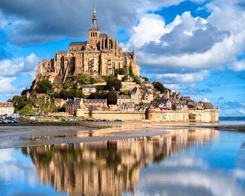 Mont Saint-Michel rises dramatically from the water, its medieval abbey and stone buildings reflected in the calm tidal surface below, under a sky of bright clouds.