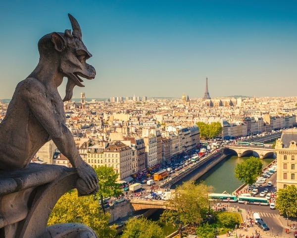 A stone gargoyle on the Notre-Dame Cathedral overlooks the city of Paris, with the Seine River, crowded streets, and the distant Eiffel Tower visible under a bright blue sky.