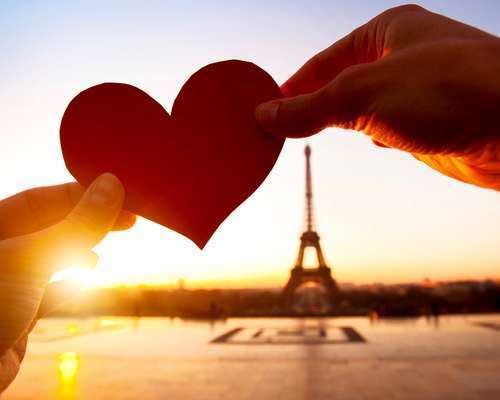 Two hands hold up a red paper heart toward the Eiffel Tower at sunrise, with warm golden light glowing behind the heart and soft silhouettes of Paris in the distance.