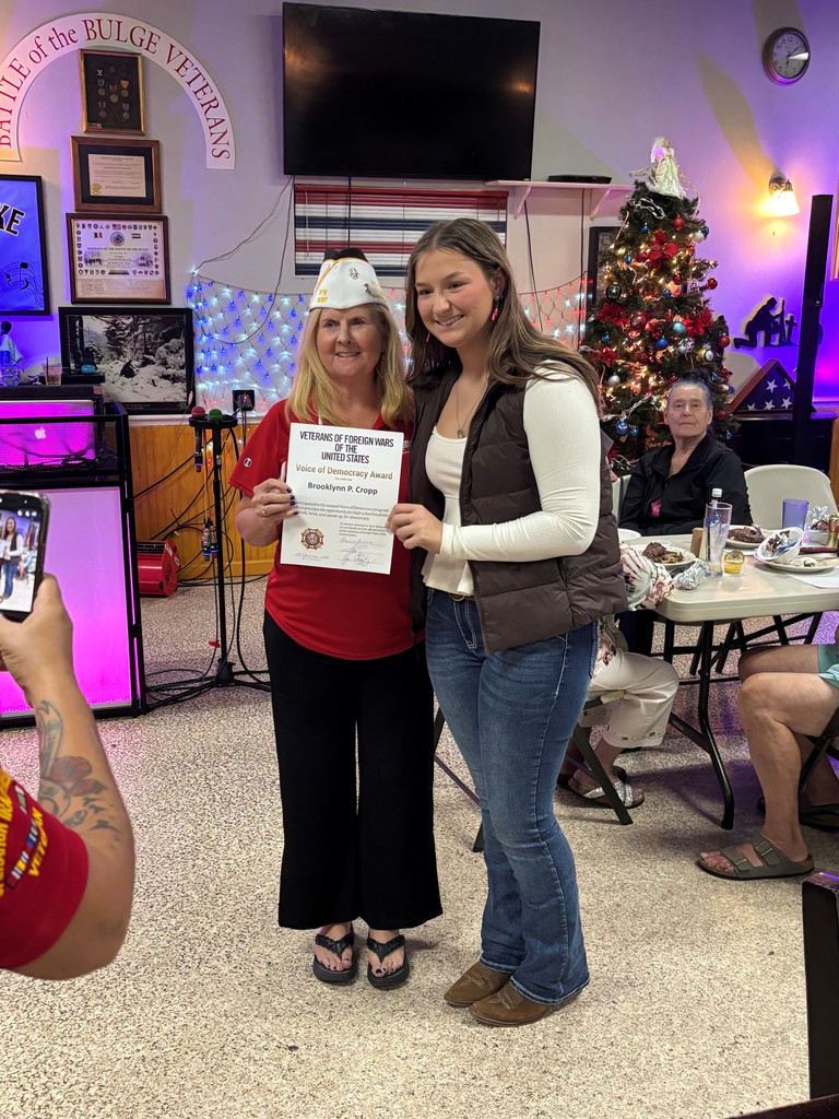 A student poses with a veteran while holding a certificate at a decorated veterans hall.