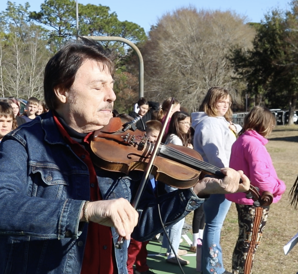 Man plays violin with eyes closed outside. STudents standing outside in line. 