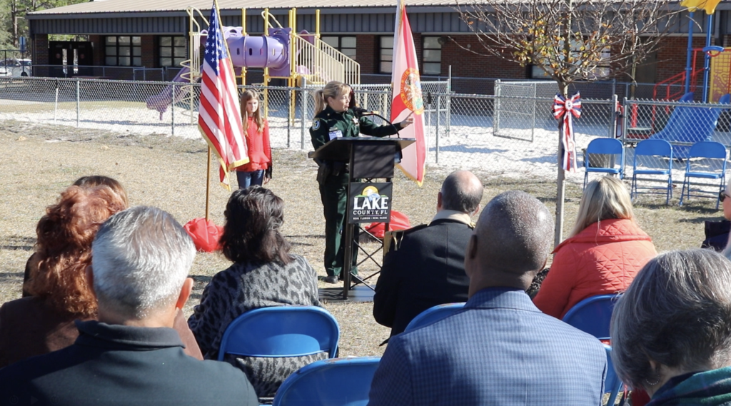 Deputy at podium outside talking to seated adult guests. She is flanked by American flag on one side, state flag on the other, and tree with red, white and blue ribbon in front