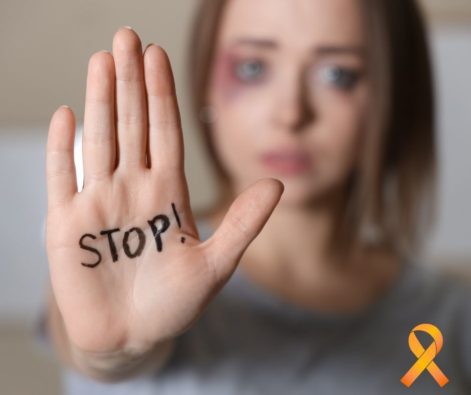 Girl with black eye holding out a hand with the word "Stop!" printed on her palm. An orange ribbon is in the bottom right corner. 