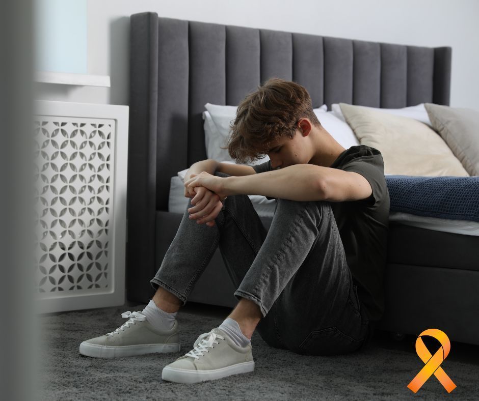 Teen boy sitting on floor by bed with head bowed looking depressed. Orange ribbon in bottom right corner.