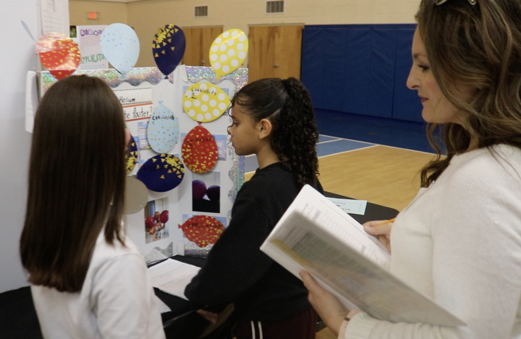 Woman judge stands behind two girls looking at their project board.